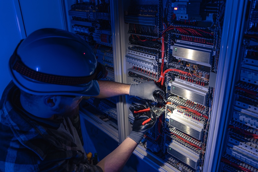 A male electrician works in a switchboard in overalls against the backdrop of emergency lighting.